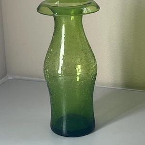 Pode incluir: Um vaso de vidro verde alto com uma textura craquelada distinta em todo o seu corpo. O vaso tem um pescoço fino e curvo que se alarga para uma borda circular na parte superior, com uma base plana e sólida para maior estabilidade.