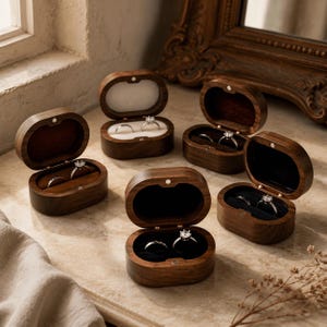 Puede incluir: Cinco cajas ovaladas para anillos de madera oscura con tapas abatibles, cada una con dos anillos de plata. Las cajas cuentan con cierres magnéticos y forros interiores de terciopelo blanco, marrón o negro, dispuestas sobre una superficie de piedra.