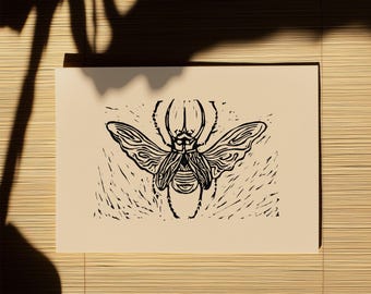 May include: A rectangular off-white paper print featuring a black ink linocut illustration of a beetle with spread wings. The insect is centered, displaying detailed line work on its body and wings, set against a textured background of short, carved marks.