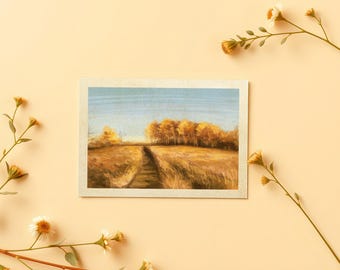 May include: A rectangular art print featuring a landscape painting of a golden field with a path leading toward a cluster of trees under a light blue sky, framed by delicate stems with small white and yellow flowers on a pale beige background.
