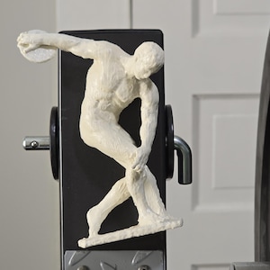 Könnte beinhalten: Eine weiße, texturierte Skulptur eines Diskuswerfers in einer dynamischen athletischen Pose, montiert auf einem schwarzen vertikalen Stützbalken. Die Figur ist in der Bewegung dargestellt, hält eine runde Scheibe und zeigt eine detaillierte Muskulatur.
