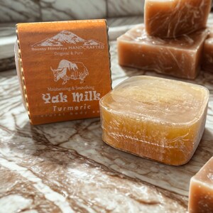 May include: A square bar of translucent orange soap wrapped in clear plastic, accompanied by a rectangular paper label featuring a mountain illustration, a yak graphic, and the text Bounty Himalaya HANDCRAFTED Original & Pure Yak Milk Turmeric.