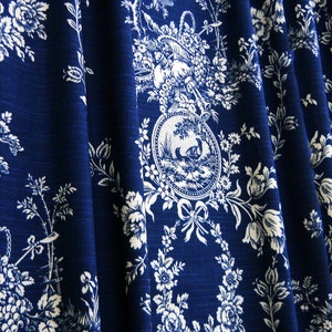Puede incluir: Tela azul oscuro con un diseño floral y pastoral blanco. El patrón incluye flores, árboles y un medallón circular con una escena de paisaje. La tela está doblada, mostrando la textura y la profundidad del material.