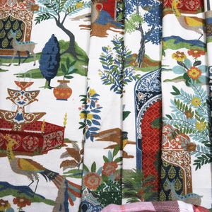 May include: White fabric with a colorful, detailed print featuring a garden scene. The design includes trees, deer, birds, and architectural elements in shades of red, blue, green, and yellow. The fabric is likely used for home decor.