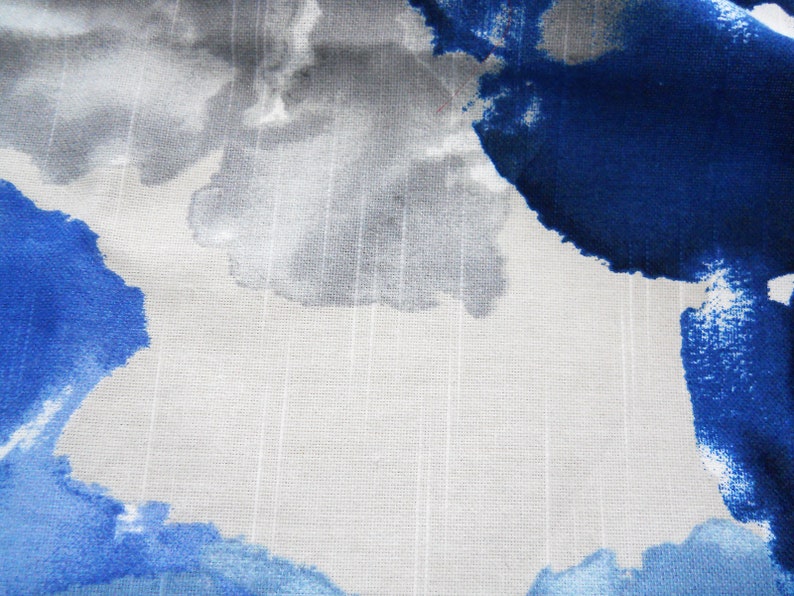 May include: Close-up of fabric with a watercolour design. The pattern features large, abstract shapes in shades of blue, grey, and white. The fabric has a textured appearance with vertical lines, creating a subtle striped effect.