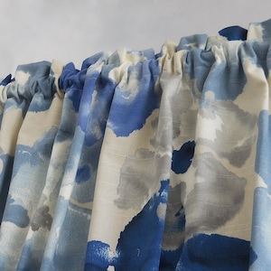 May include: Curtains with a blue and white watercolour design, hanging from a gold-coloured rod. The fabric has a pattern of blue and grey brushstrokes on a white backdrop. The rod has decorative finials.
