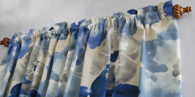 May include: Curtains with a blue and white watercolour design, hanging from a gold-coloured rod. The fabric has a pattern of blue and grey brushstrokes on a white backdrop. The rod has decorative finials.