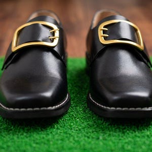 May include: A pair of black leather shoes featuring a square toe design and a prominent gold-toned metal buckle across the top strap. The shoes have visible white stitching along the sole edge and a smooth, polished finish on the upper surface.