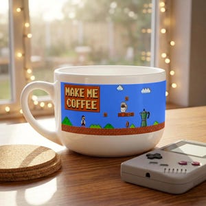 Op de afbeelding: Een witte keramische mok met een blauwe pixel-videogame landschapsafbeelding. Het ontwerp bevat een bruin bord met de tekst "MAKE ME COFFEE", een klein personage, een koffiepot en bruine bakstenen platforms tegen een helderblauwe lucht.