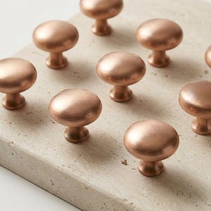 May include: A collection of round cabinet knobs with a brushed copper finish. Each knob features a smooth, domed top and a tapered base, arranged in a grid pattern. The metallic surface reflects light, highlighting the warm, reddish-brown tone.