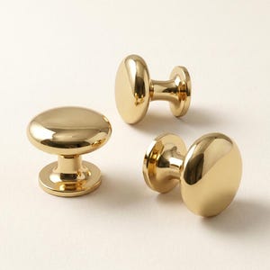 May include: Three polished gold-tone cabinet knobs with a smooth, rounded mushroom-shaped head and a cylindrical base. Each knob features a reflective metallic finish and a circular mounting plate, designed for attachment to furniture or cabinetry.