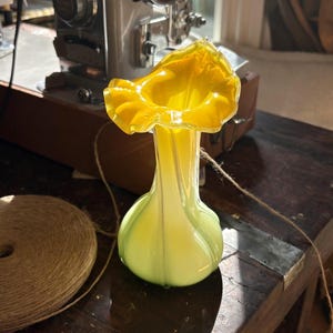 May include: A glass vase featuring a bulbous base that transitions from a pale yellow-green at the bottom to a vibrant, saturated yellow at the top. The rim is wide and ruffled, creating a flared opening with a distinct, wavy, and scalloped edge.