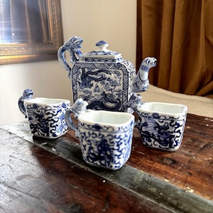 May include: A white porcelain tea set featuring blue hand-painted dragon motifs. The set includes a square teapot with a lid and three matching square cups. Each piece is adorned with intricate blue patterns and a sculpted dragon handle.