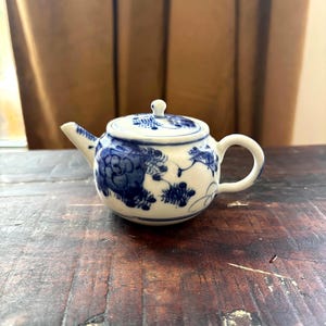 May include: A small white ceramic teapot featuring a blue floral pattern painted on the exterior. The teapot has a rounded body, a curved handle, a short spout, and a matching lid with a small knob handle on top, all decorated in blue.
