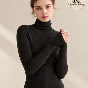 May include: A dark charcoal gray long-sleeve turtleneck sweater with a fitted silhouette. The garment features a ribbed knit texture and a high neck. A circular logo in the upper right corner displays the text Ravelle Clothing.