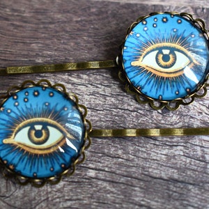K&ouml;nnte beinhalten: Zwei Haarspangen mit runden, bronzefarbenen Fassungen und gewelltem Rand. Jede Spange enth&auml;lt einen Glaskabochon mit der Grafik eines stilisierten Auges mit brauner Iris, umgeben von goldenen Strahlen und kleinen Sternen auf blauem Grund.