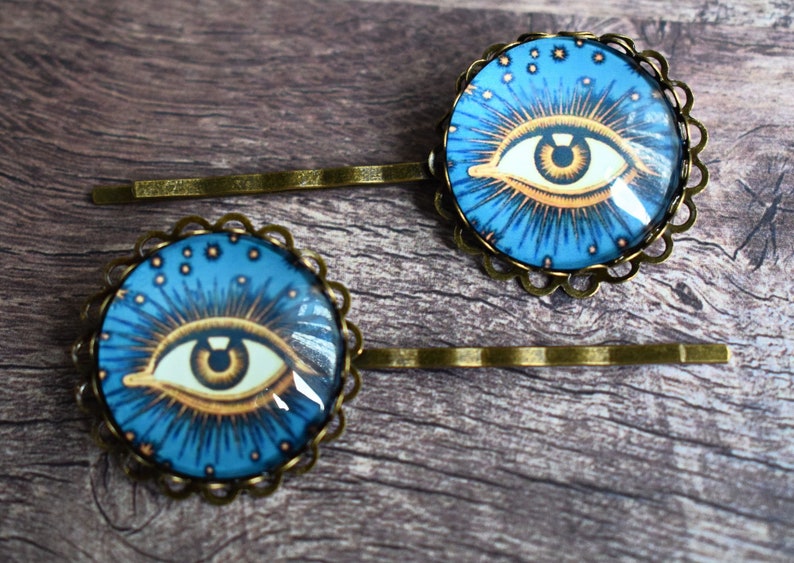 K&ouml;nnte beinhalten: Zwei Haarspangen mit runden, bronzefarbenen Fassungen und gewelltem Rand. Jede Spange enth&auml;lt einen Glaskabochon mit der Grafik eines stilisierten Auges mit brauner Iris, umgeben von goldenen Strahlen und kleinen Sternen auf blauem Grund.
