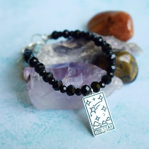 May include: Black beaded bracelet with a silver charm featuring the The Star tarot card illustration. The charm has a white background with a black outline.