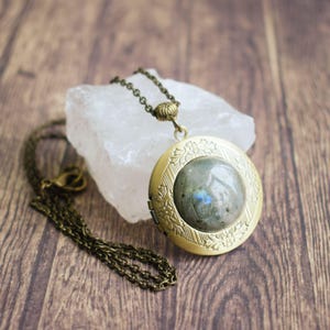 May include: A brass locket necklace featuring a round labradorite gemstone pendant. The locket has an ornate floral design around the edge.