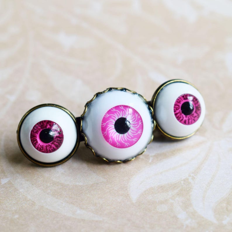 K&ouml;nnte beinhalten: Eine Haarspange mit drei runden, wei&szlig;en, augenf&ouml;rmigen Cabochons in antik bronzefarbenen Metallfassungen. Jedes Auge hat eine leuchtend pinke Iris mit feinen radialen Linien und eine tiefschwarze Pupille, was ein auff&auml;lliges Accessoire ergibt.