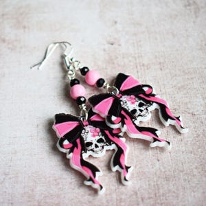 May include: Pink and black skull earrings with a bow design. The earrings are made of polymer clay and have silver hooks.
