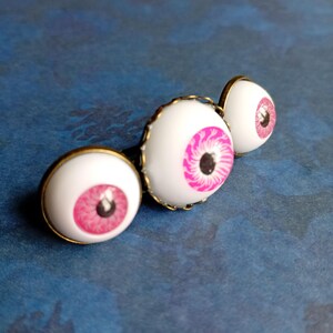 K&ouml;nnte beinhalten: Ein Set aus drei runden Cabochon-Anstecknadeln mit realistischen wei&szlig;en Aug&auml;pfeln, leuchtend pinken Iris und schwarzen Pupillen, die jeweils in einer schlichten, antik-bronzefarbenen Metallfassung sitzen und in einer Reihe angeordnet sind.