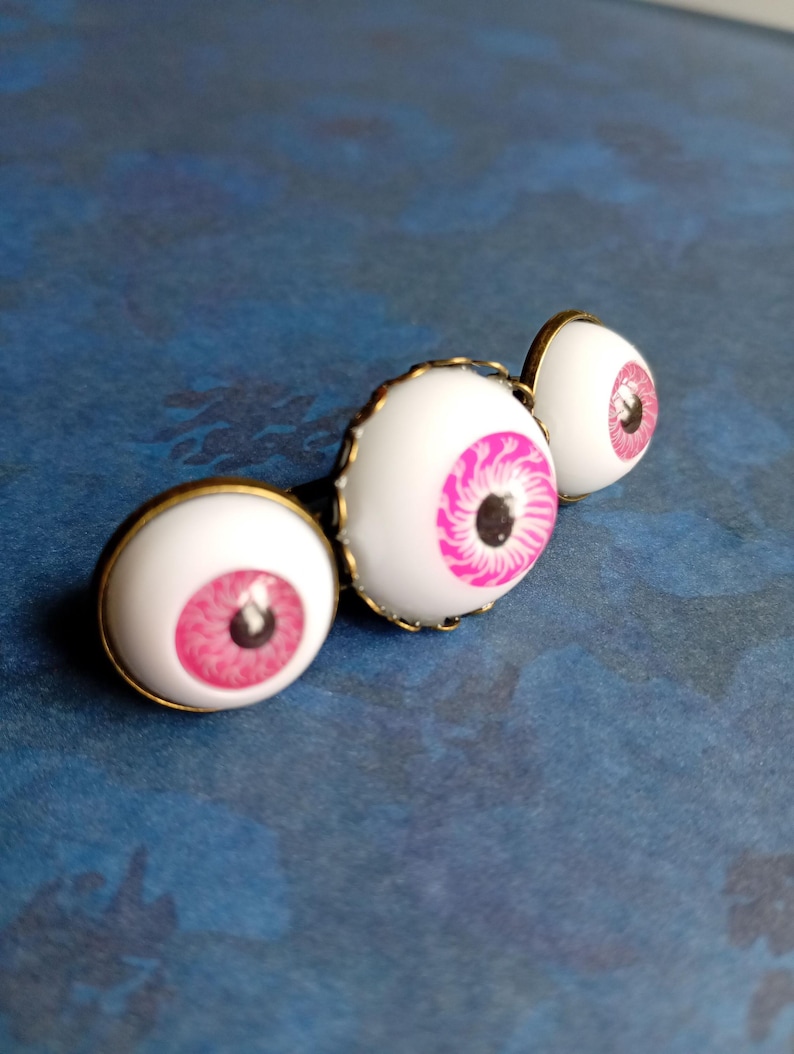 K&ouml;nnte beinhalten: Ein Set aus drei runden Cabochon-Anstecknadeln mit realistischen wei&szlig;en Aug&auml;pfeln, leuchtend pinken Iris und schwarzen Pupillen, die jeweils in einer schlichten, antik-bronzefarbenen Metallfassung sitzen und in einer Reihe angeordnet sind.