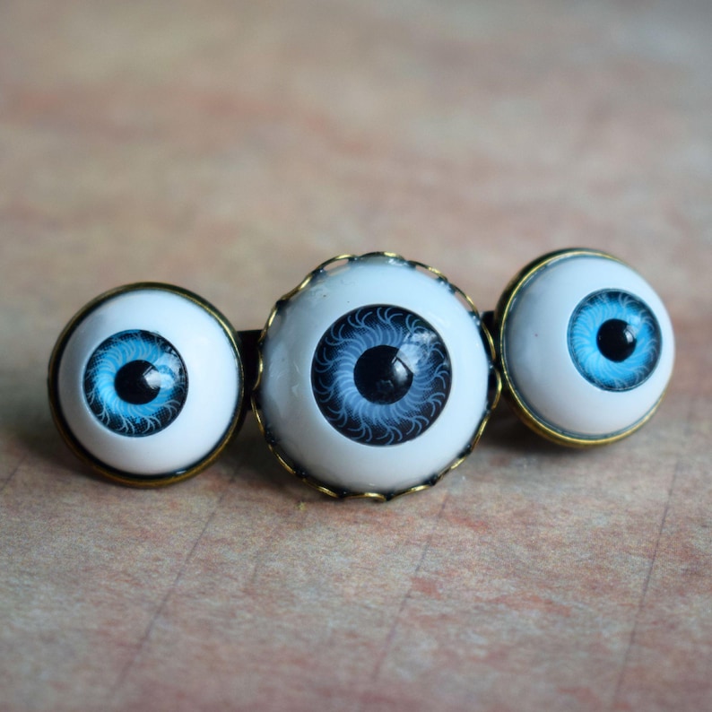 K&ouml;nnte beinhalten: Ein Haaraccessoire mit drei runden, realistischen Augencabochons mit leuchtend blauen Iris und schwarzen Pupillen. Jedes Auge ist in einen metallischen, bronzefarbenen Rahmen gefasst, wobei das mittlere Auge etwas gr&ouml;&szlig;er als die beiden &auml;u&szlig;eren ist.