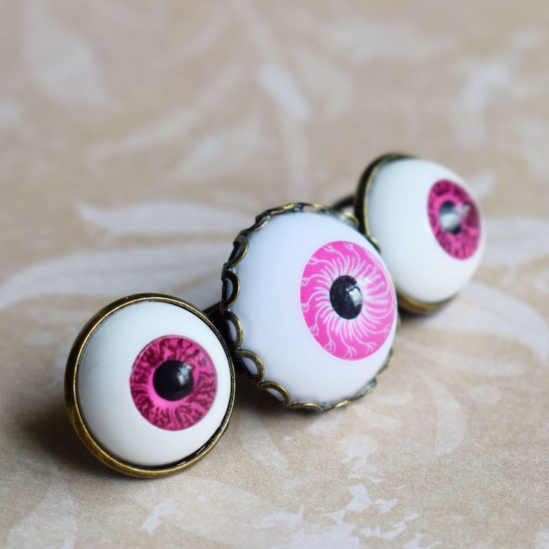 K&ouml;nnte beinhalten: Drei runde Cabochon-Kn&ouml;pfe mit einem realistischen Augendesign, bestehend aus einer wei&szlig;en Sklera, einer leuchtend pinken Iris mit detaillierten radialen Linien und einer schwarzen Pupille. Jeder Knopf ist in einem gewellten, bronzefarbenen Metallrahmen gefasst.