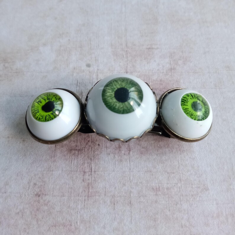 K&ouml;nnte beinhalten: Eine Haarspange mit drei realistischen, kugelf&ouml;rmigen gr&uuml;nen Augencabochons in bronzefarbenen Metallfassungen. Das mittlere Auge ist gr&ouml;&szlig;er als die beiden seitlichen Augen, wobei jedes eine detaillierte gr&uuml;ne Iris und eine schwarze Pupille zeigt.