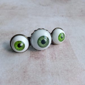K&ouml;nnte beinhalten: Ein Haaraccessoire mit drei runden, realistisch wirkenden gr&uuml;nen Augen mit schwarzen Pupillen, die in bronzefarbene Metallfassungen eingefasst sind. Die Augen sind in einer Reihe angeordnet, wobei das mittlere Auge etwas gr&ouml;&szlig;er ist.