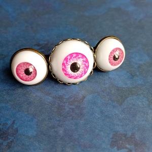 K&ouml;nnte beinhalten: Ein Haaraccessoire mit drei runden, wei&szlig;en Augapfel-Cabochons in bronzefarbenen Metallfassungen. Jeder Augapfel hat eine leuchtend pinke Iris mit feinen schwarzen radialen Linien und einer zentralen schwarzen Pupille, horizontal angeordnet.