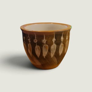 Può includere: Una piccola tazza in ceramica arrotondata con esterno marrone e interno bianco. La superficie esterna presenta un motivo dipinto a mano di forme bianche a goccia che pendono da sottili linee verticali attorno al vaso.