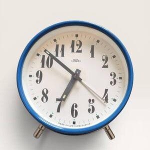 May include: A round analog alarm clock with a bright blue metal frame and a white face. The clock features black Arabic numerals, black hour and minute hands, a thin silver second hand, and the text "MADE IN CZECHOSLOVAKIA" at the bottom.