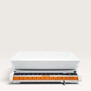 May include: A white mechanical kitchen scale featuring a rectangular weighing platform and a horizontal display panel. The scale has an orange measurement bar with black text indicating a capacity of up to 13 kilograms and 28 pounds.