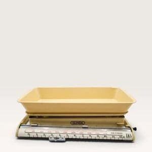 May include: A vintage Gunda kitchen scale featuring a pale yellow rectangular metal tray resting on a matching yellow base. The front displays a horizontal sliding weight mechanism with a clear scale marked in increments of 100 grams up to 10 kg.