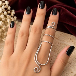 Puede incluir: Un anillo de armadura para dedo en metal plateado con un diseño largo y vertical que cubre la longitud del dedo. La pieza consta de dos bandas finas y curvas en la parte superior y un marco ovalado alargado en la base con extremos en espiral.