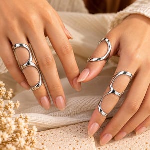Puede incluir: Tres anillos de metal plateado con un diseño alargado de marco abierto que cubre dos articulaciones de los dedos. Cada anillo consta de dos bandas circulares conectadas por una barra vertical, creando una estética elegante y pulida.