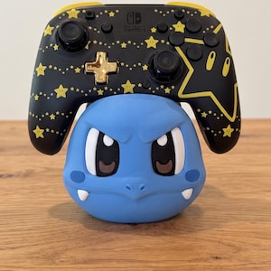 May include: A black Nintendo Switch Pro controller featuring a yellow star pattern and a gold D-pad, resting on a light blue, round, character-shaped stand with large eyes, a small nose, and two white fangs protruding from the bottom.