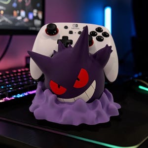 May include: A purple Gengar character figure serving as a controller stand, featuring red eyes and a wide grin, holding a white Nintendo Switch Pro controller with black buttons and red accents on the analog sticks, set on a light purple base.