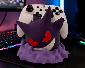 May include: A purple Gengar character figure serving as a controller stand, featuring red eyes and a wide grin, holding a white Nintendo Switch Pro controller with black buttons and red accents on the analog sticks, set on a light purple base.