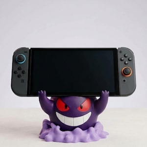 May include: A purple Gengar character figure acting as a stand for a black handheld gaming console. The figure has red eyes, a wide white grin, and raised arms holding the device. The console features grey controllers with blue and orange accents.