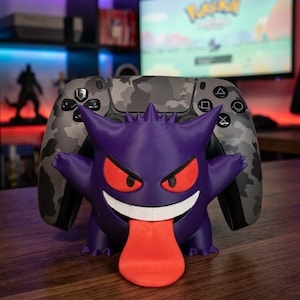 May include: A purple Gengar character controller stand featuring red eyes, a white grin, and a long red tongue, holding a gray and black camouflage patterned video game controller with black buttons and directional pads.