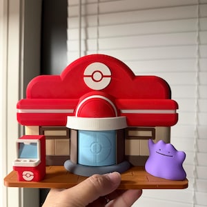 May include: A 3D-printed model of a red and white Pokemon Center building featuring a central blue door, a small red terminal on the left, and a purple Ditto figure on the right, all mounted on a brown rectangular base with a flat surface.
