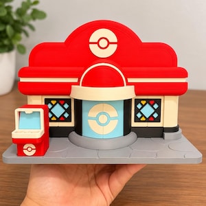 May include: A red and cream-colored plastic model of a building featuring a central light blue door with a circular emblem, two side windows with geometric patterns, a red roof with a circular emblem, and a small red arcade machine on the side.