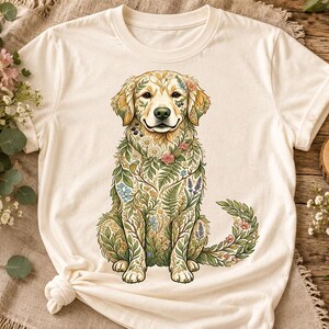 Może przedstawiać: Kremowy t-shirt z krótkim rękawem z centralną grafiką przedstawiającą psa rasy golden retriever. Sierść psa jest ozdobiona misternym wzorem składającym się z różnorodnych ilustracji botanicznych, w tym zielonych liści, paproci i kwiatów.