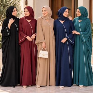 Puede incluir: Cinco abayas de manga larga en negro, burdeos, beige, azul marino y verde azulado, cada una con un hiyab a juego y un ribete diagonal blanco en la parte delantera. La abaya beige se combina con un pequeño bolso blanco estructurado con herrajes dorados.