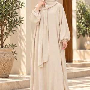 Puede incluir: Un vestido abaya beige de cuerpo entero con mangas largas y holgadas con puños elásticos fruncidos y un pañuelo hijab beige a juego. La prenda tiene una silueta sencilla y fluida con un cuello alto y un color neutro y sólido en toda la pieza.