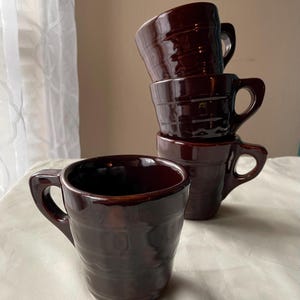 May include: A set of four dark brown ceramic mugs with a glossy finish and a textured, ribbed exterior. Each mug features a sturdy handle and a tapered shape. One mug stands alone in the foreground, while three others are stacked vertically behind it.