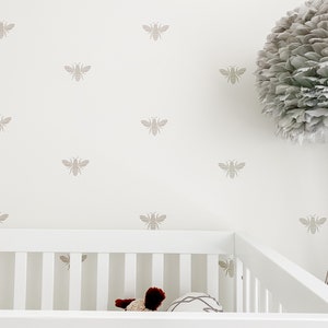 May include: A white cot with a white wooden frame and a white mattress. The wall behind the cot is white with a pattern of beige bees. A large grey feather lamp sits on a stand to the right of the cot.